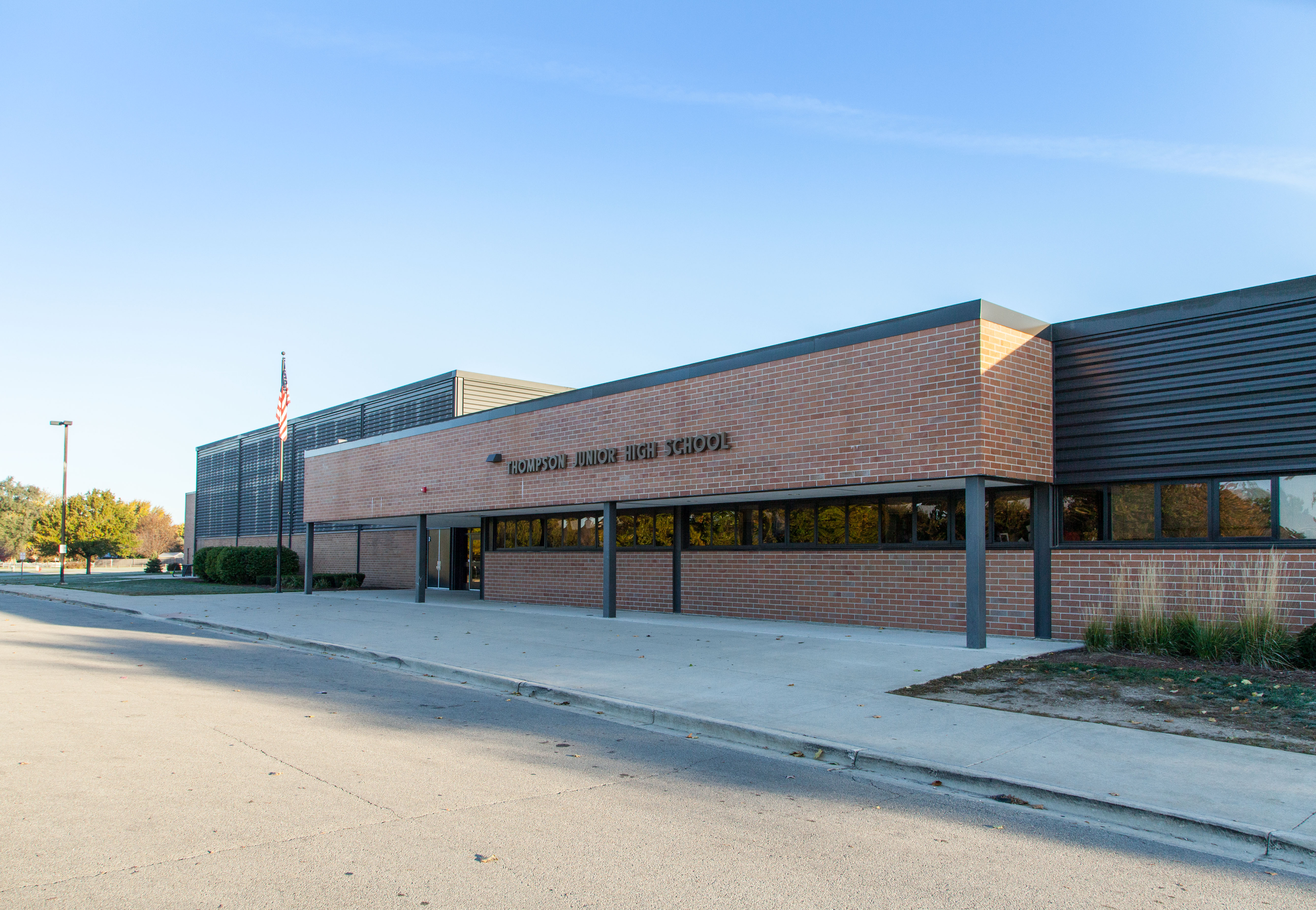 Building front of Thompson showing long curb, building entrance, American flag, and blue sky in background. 